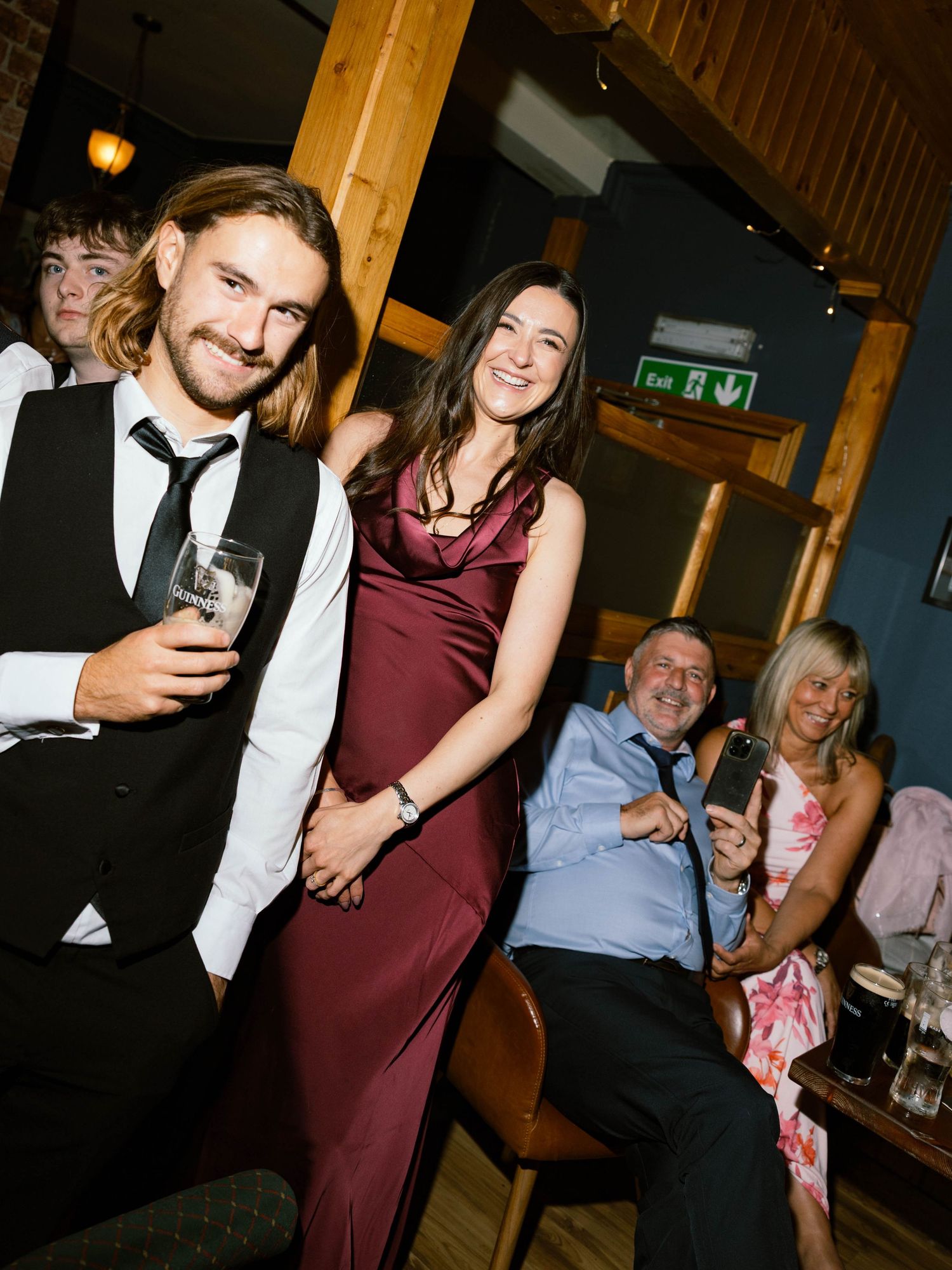 Party guests in formal attire enjoying drinks at a social gathering with a burgundy dress and tuxedo vest visible.
