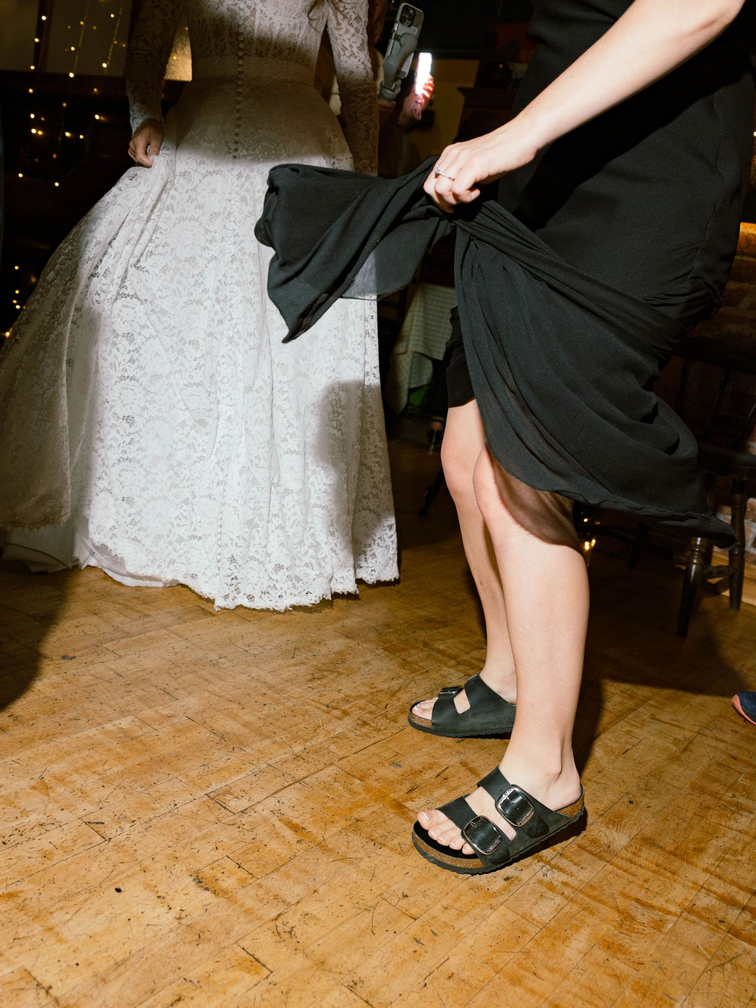 Person in black clothes and sandals stands next to a white lace wedding dress on a wooden dance floor.