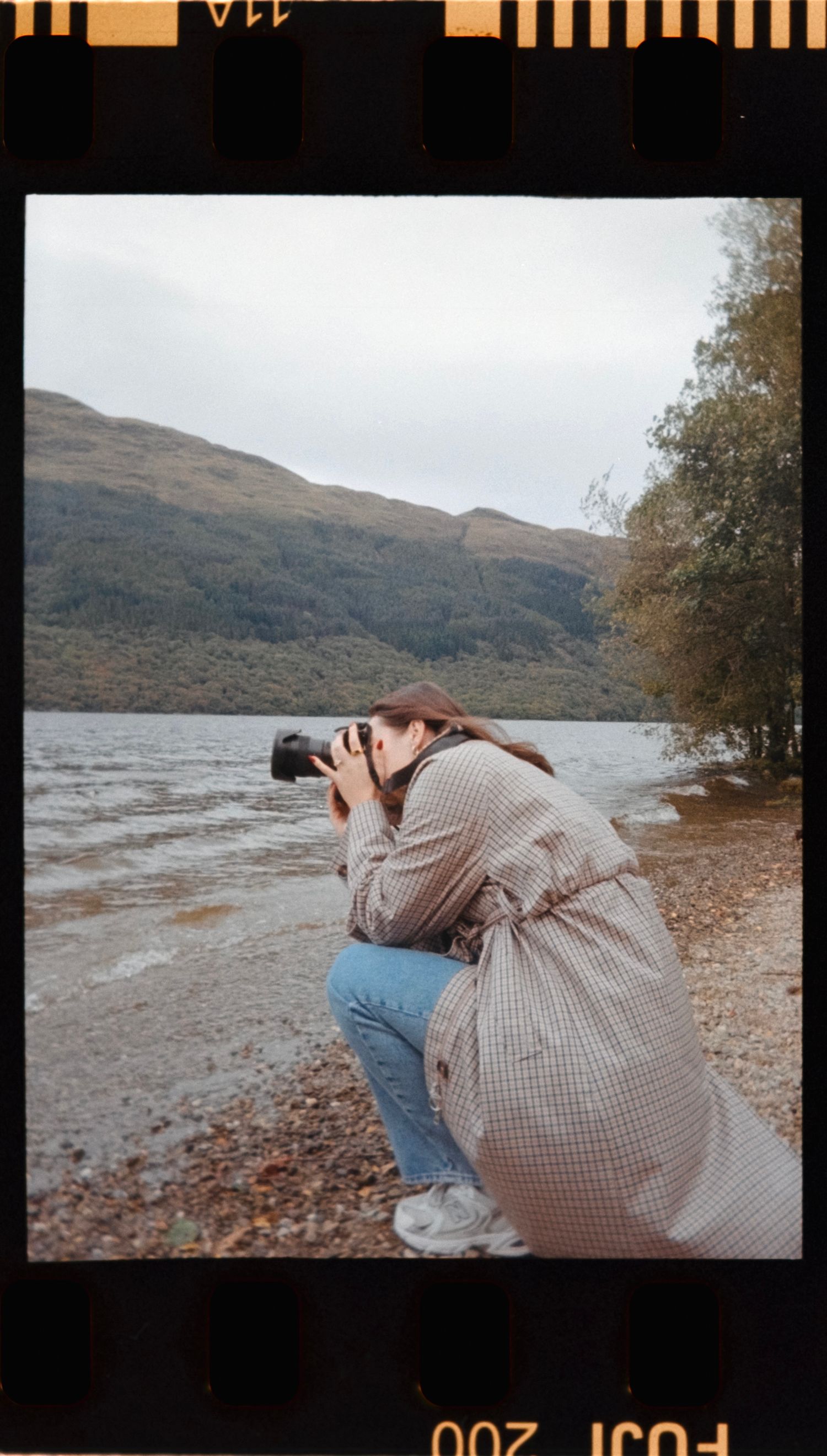 A photographer in a plaid coat takes pictures by a scenic lake and mountains.