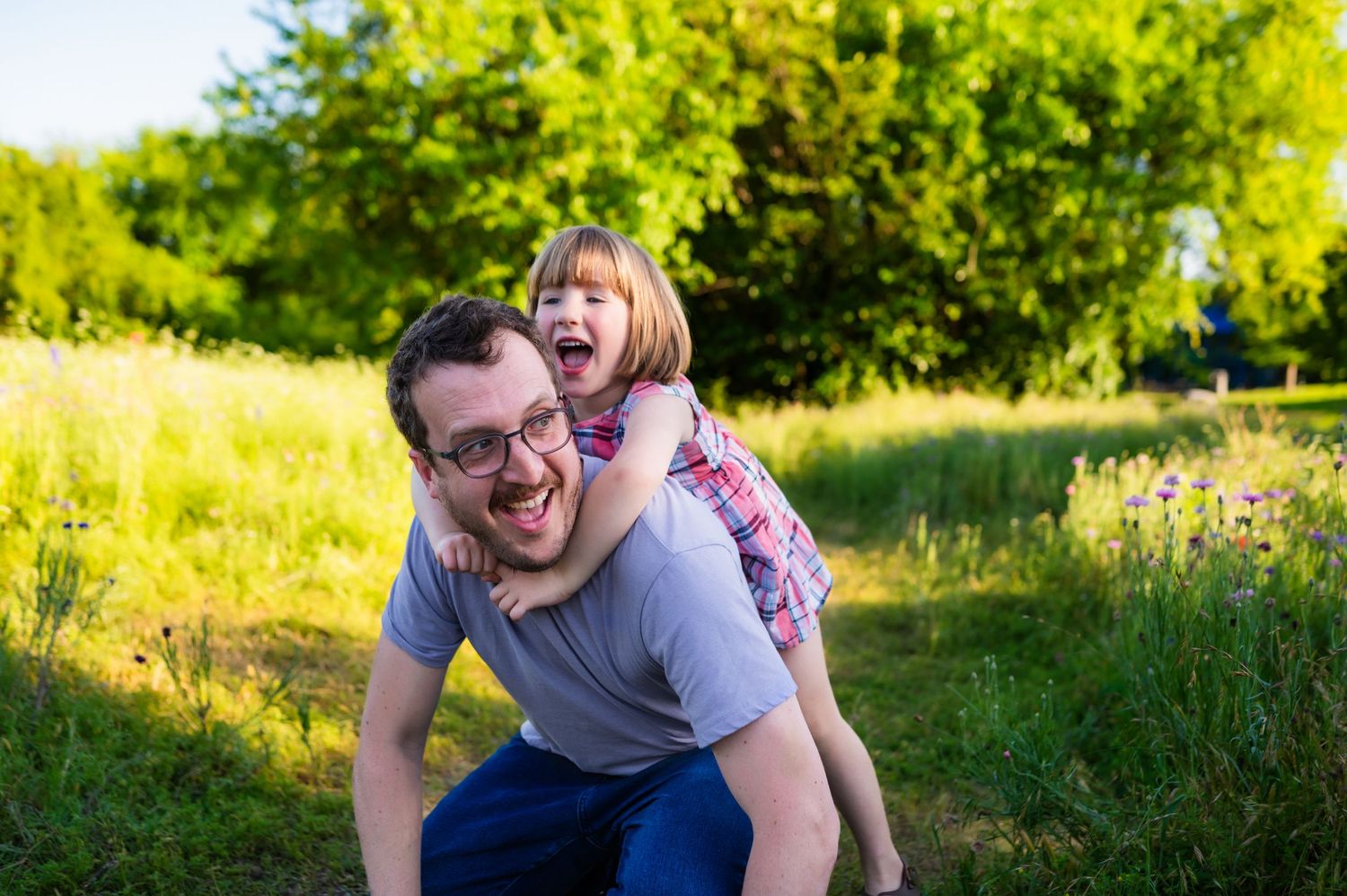 Richardson Dad kneeling during outdoor family mini session smiling looking back w/ preschool aged daughter laughing and climbing on his back
