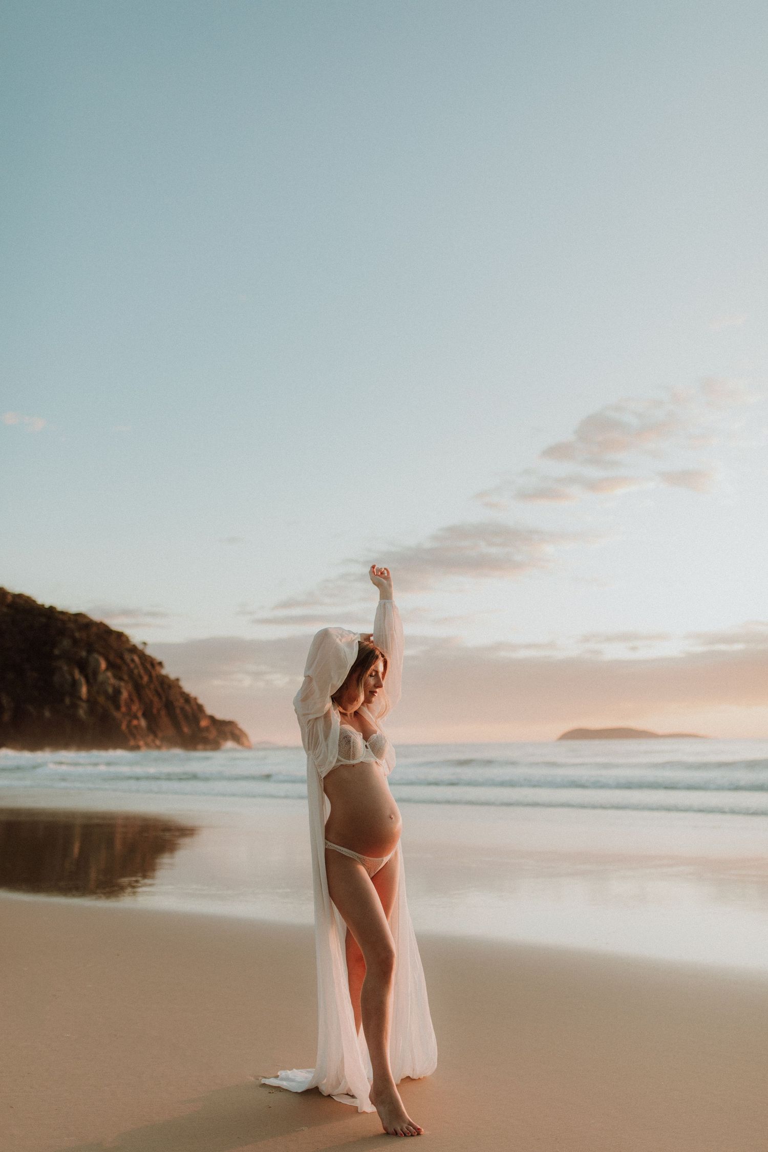 A person stands on a sandy beach at sunset wearing white fabric that flows in the ocean breeze.