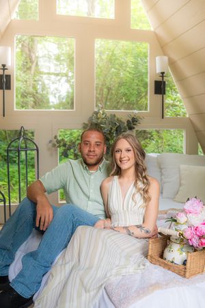 A couple relaxes together on a white couch in a bright sunlit room with windows and pink flowers.