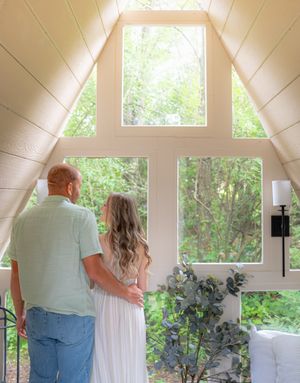 A couple stands together in a bright white room with large windows and indoor plants.