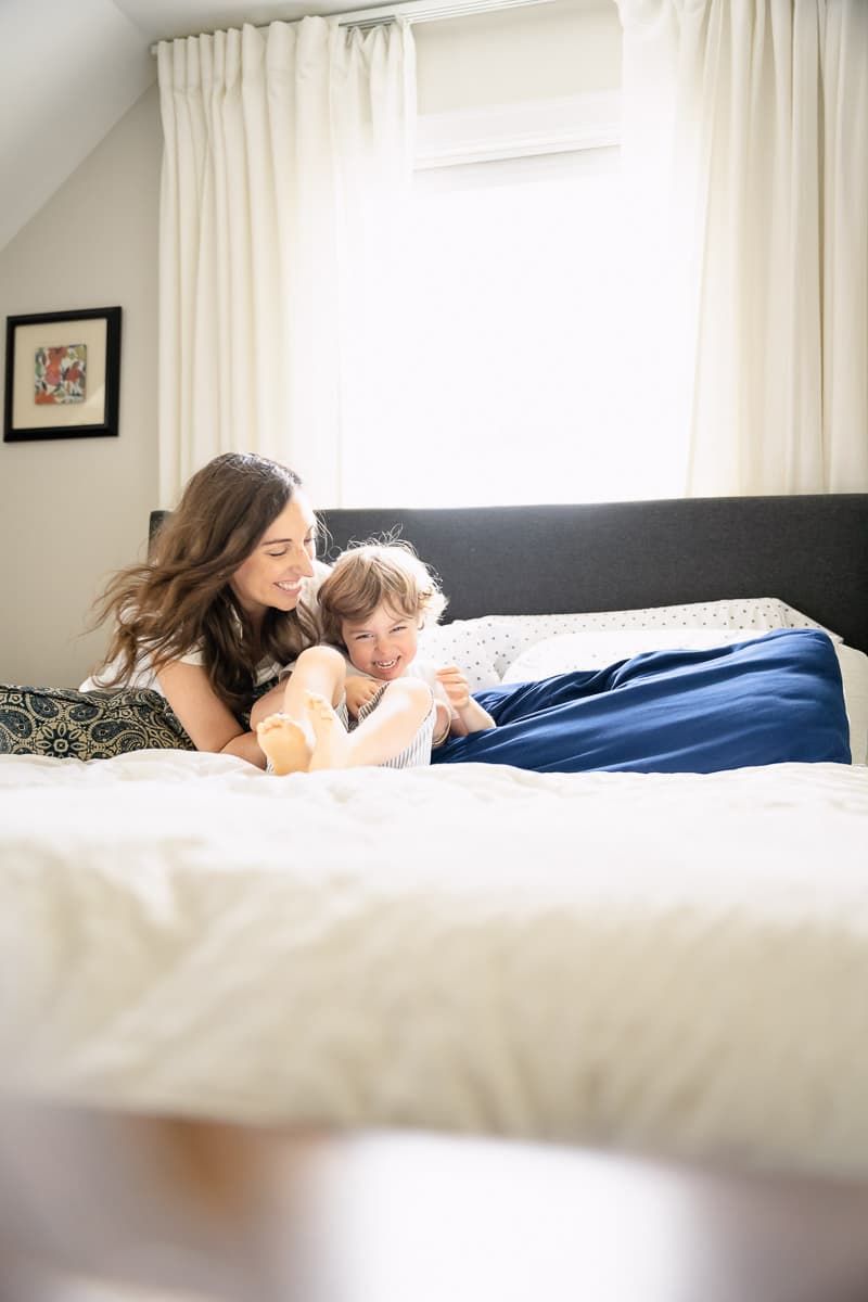 A heartwarming moment shared between mom and son relaxing together on a white bed with dark headboard in a bright bedroom.
