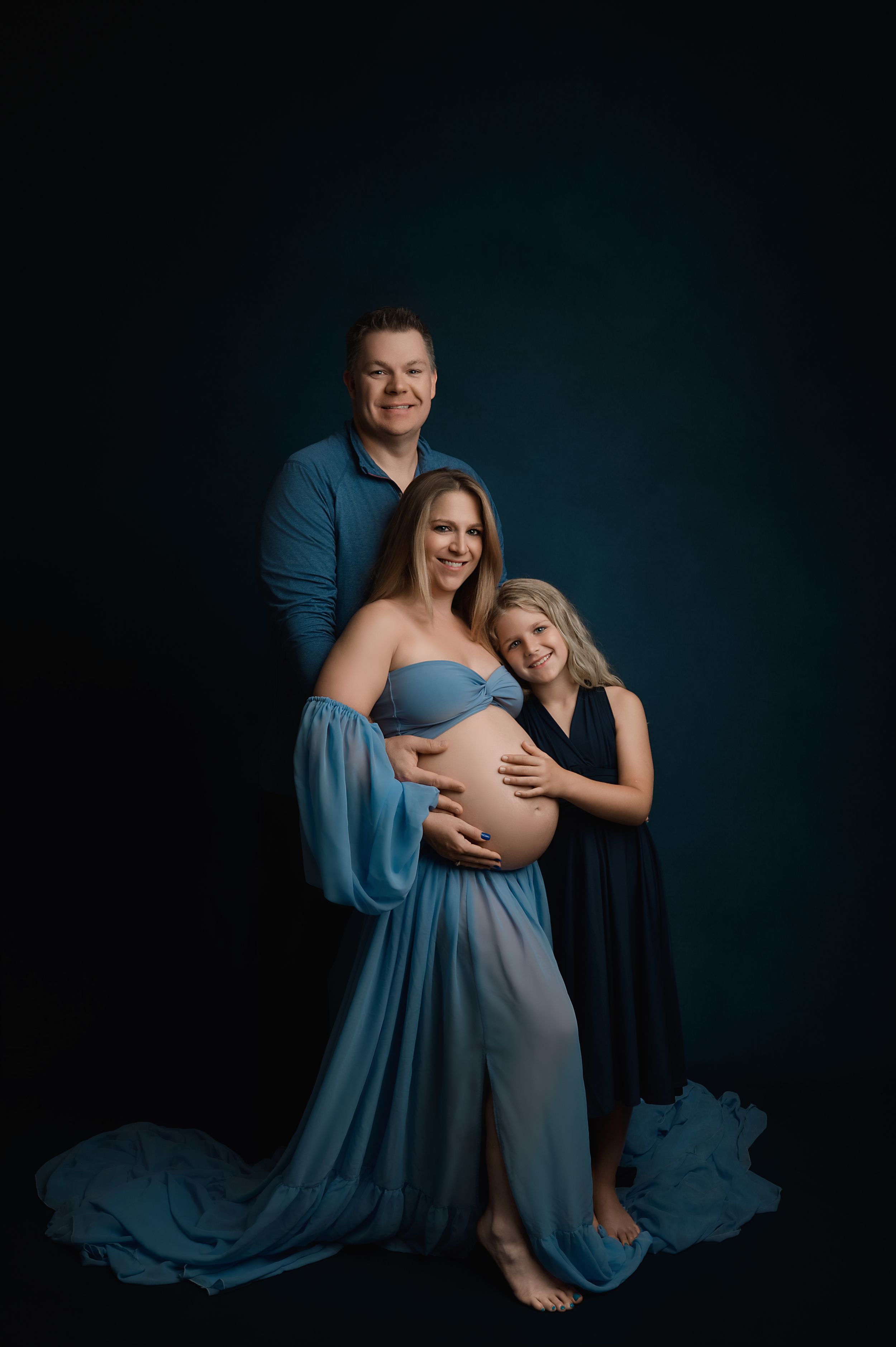 Intimate maternity photoshoot shows couple embracing in blue dress against dark backdrop with artistic lighting and poses.