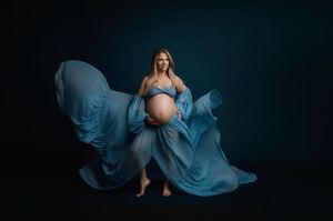 A maternity photo featuring flowing light blue fabric dramatically billowing around an expectant mother against a dark background.