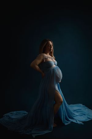 A dramatic maternity portrait features an expectant mother in a flowing blue gown against a dark moody backdrop.