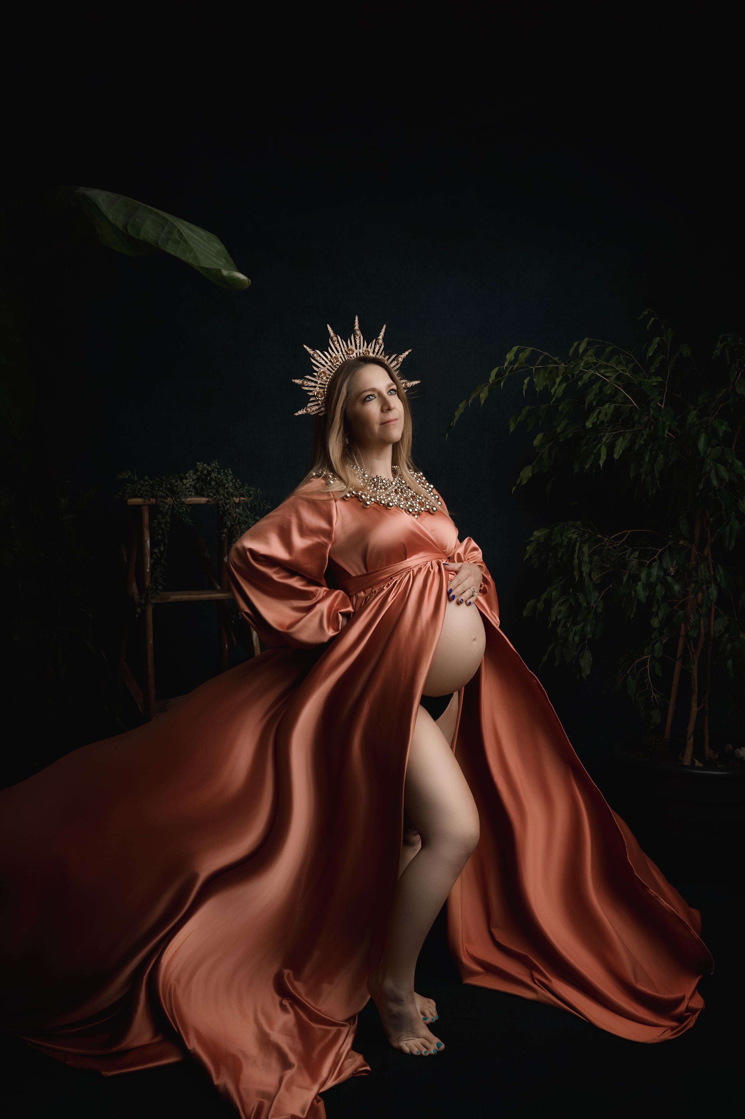 Dramatic artistic maternity photoshoot with a person in flowing copper silk gown and crown against dark background.
