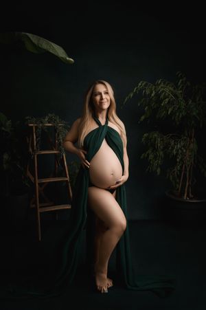 A maternity photo showing an expectant mother in an elegant emerald green draped fabric against a dark moody backdrop.