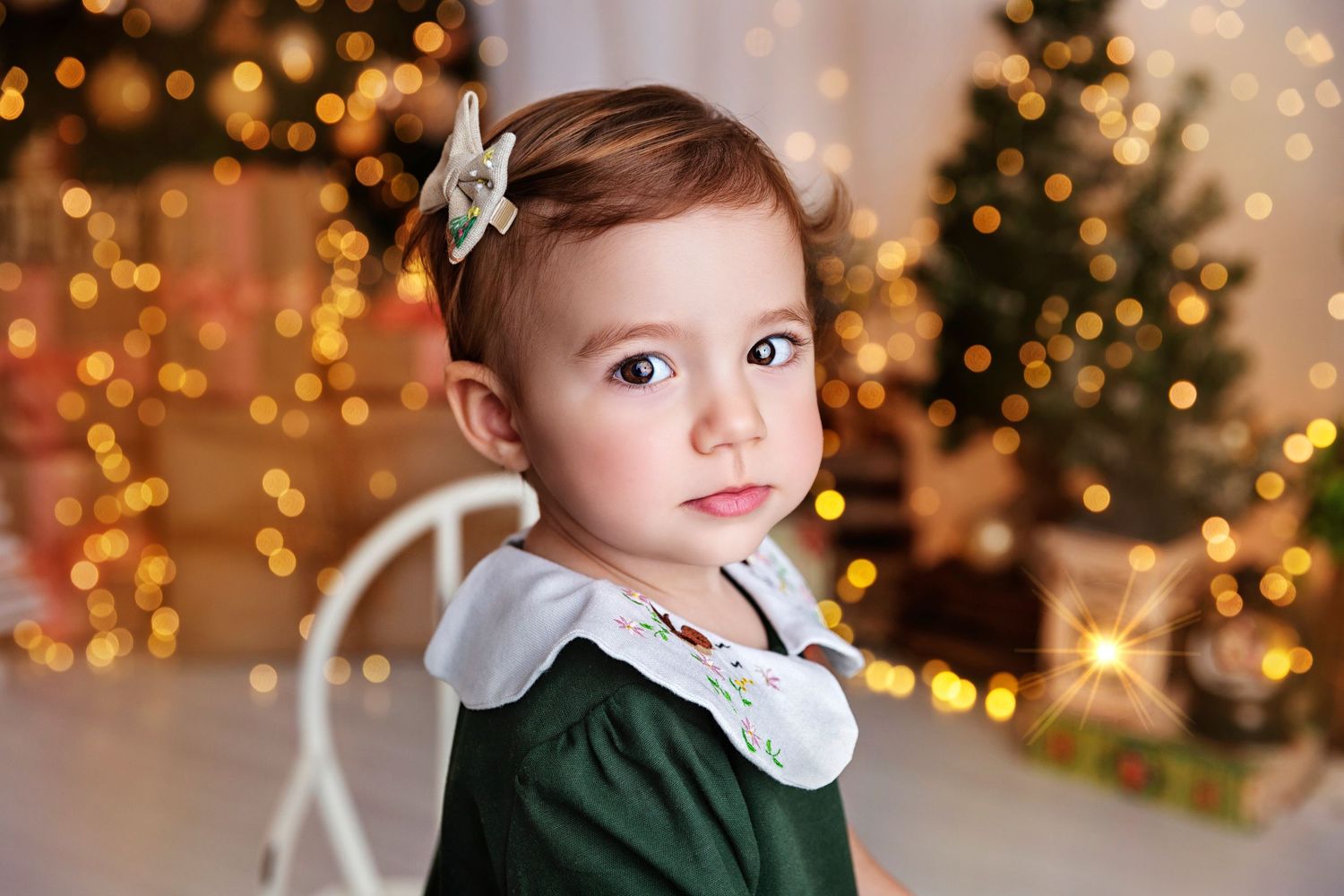 A small child in a green holiday dress with a white collar poses in front of a glowing Christmas tree backdrop.