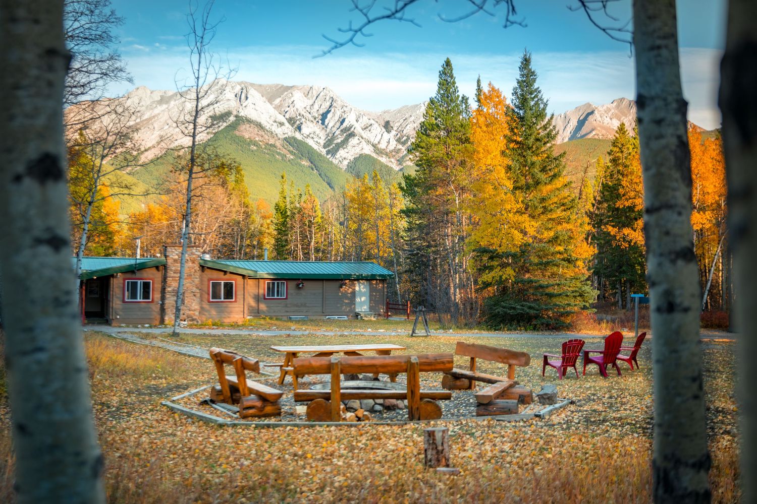 Canadian flag waves over a rustic wooden cabin with green roof surrounded by autumn trees against blue sky.