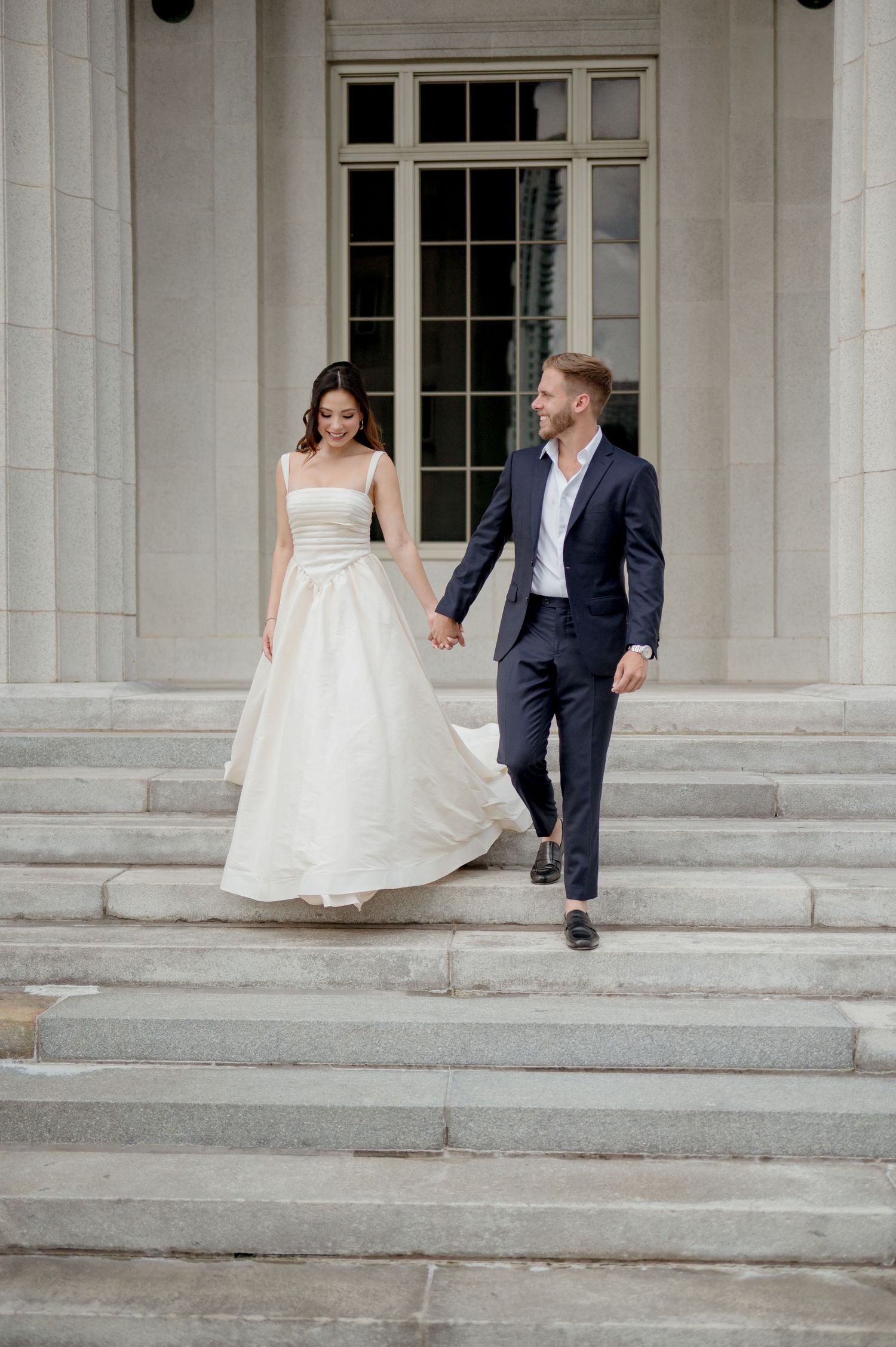 A couple descending grand stone steps, with the bride wearing a white gown and the groom in a navy suit.