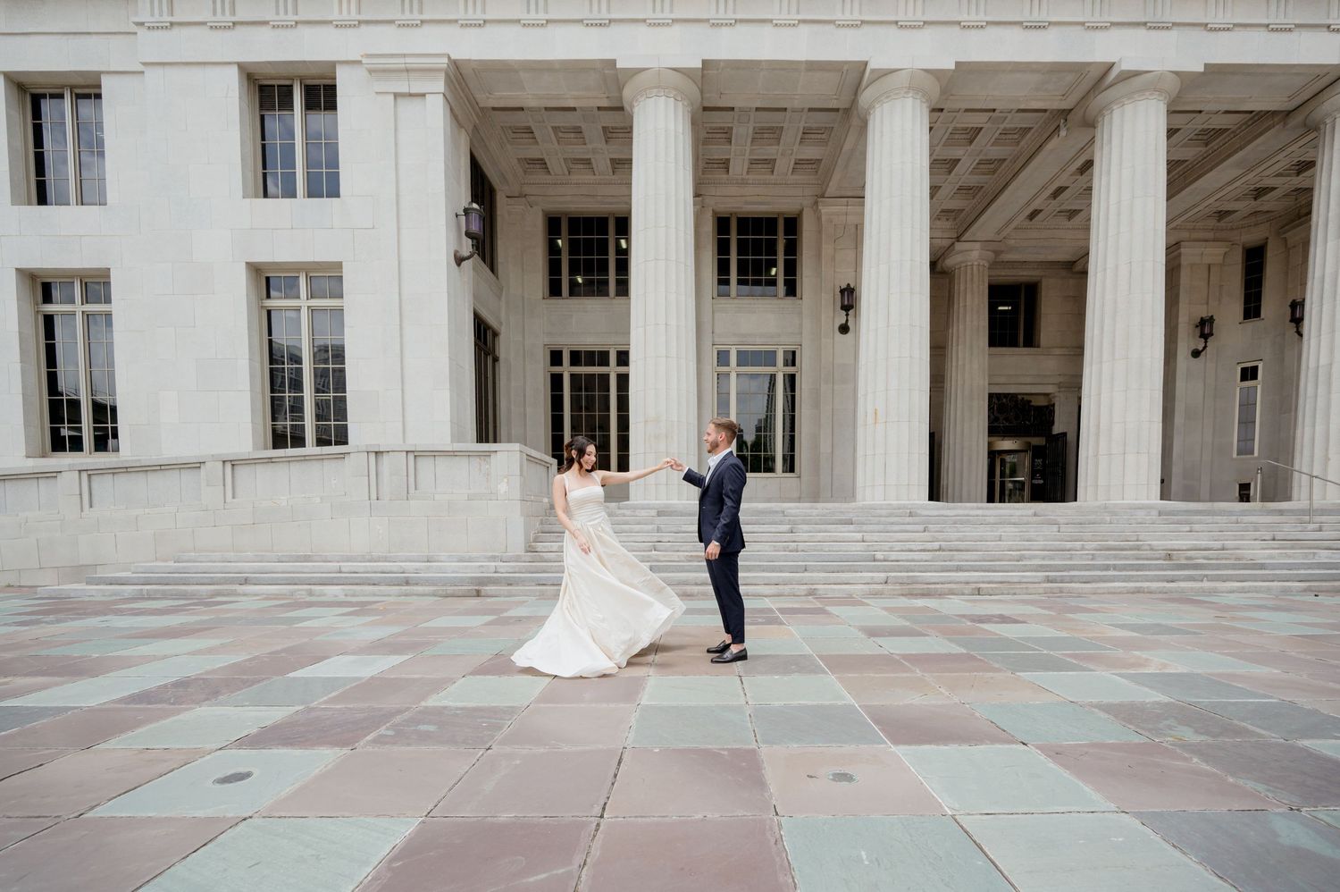 A bride and groom pose for wedding photos in front of a grand white marble colonnade building with classical architecture.