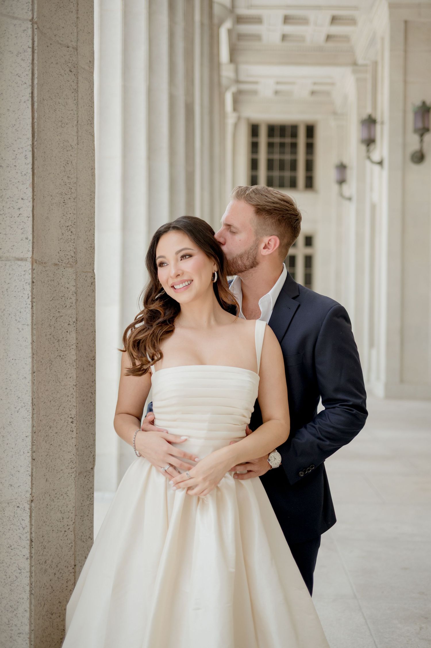Elegant couple poses together in formal wedding attire underneath a beautiful columned archway.