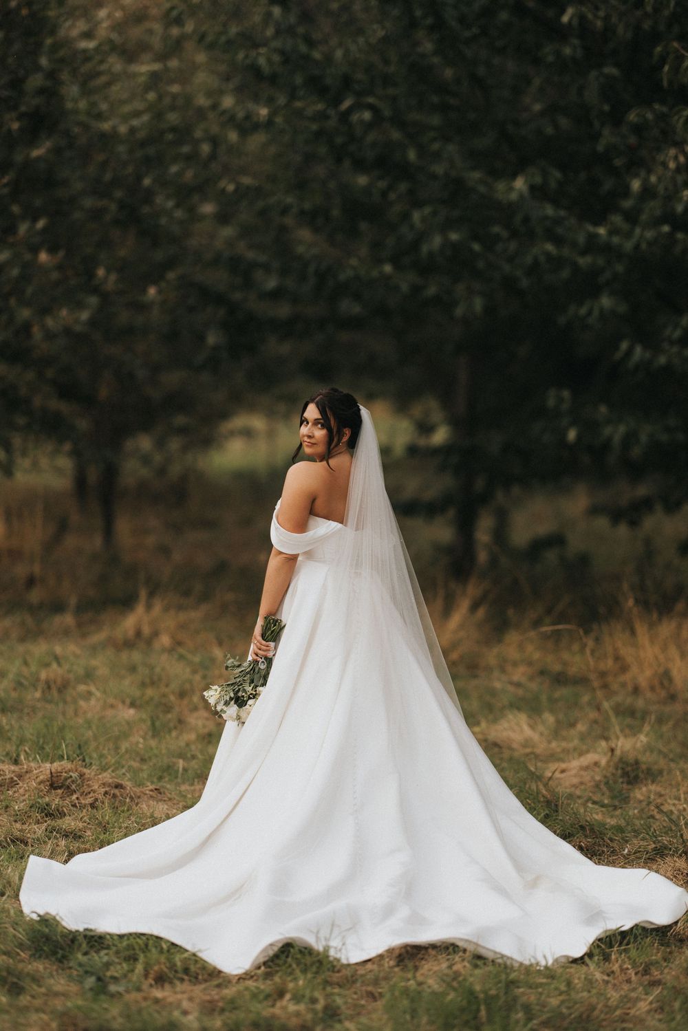 A bride in an elegant off-shoulder white wedding gown with long flowing veil poses against a rustic outdoor backdrop.