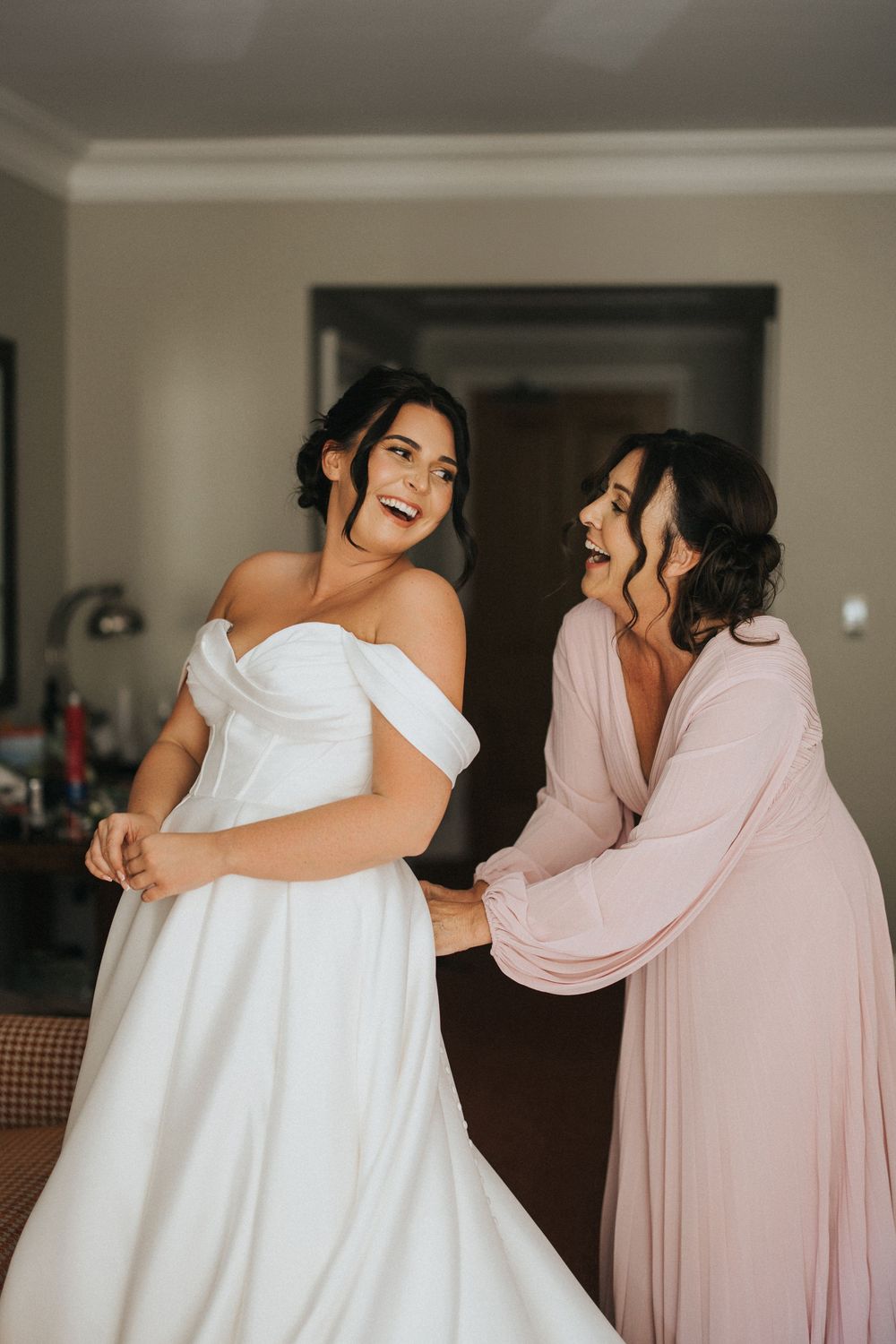 A bride in white off-shoulder wedding gown shares a joyful moment with bridesmaid in pink flowing dress during preparations.