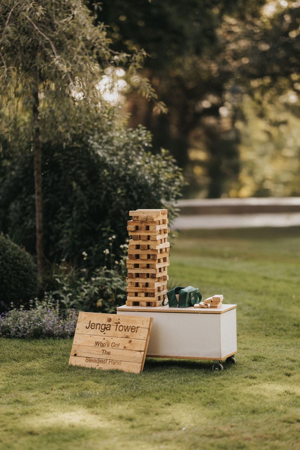 Outdoor Jenga tower game setup with wooden blocks and sign on grassy lawn surrounded by trees.