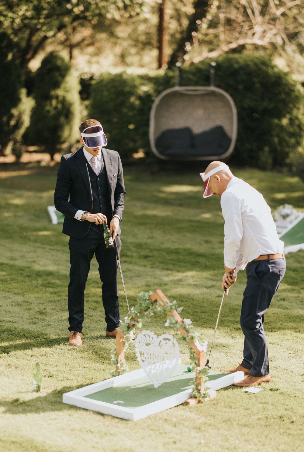 Two golfers practice putting on a mini golf course setup in a sunny garden area with decorative elements.
