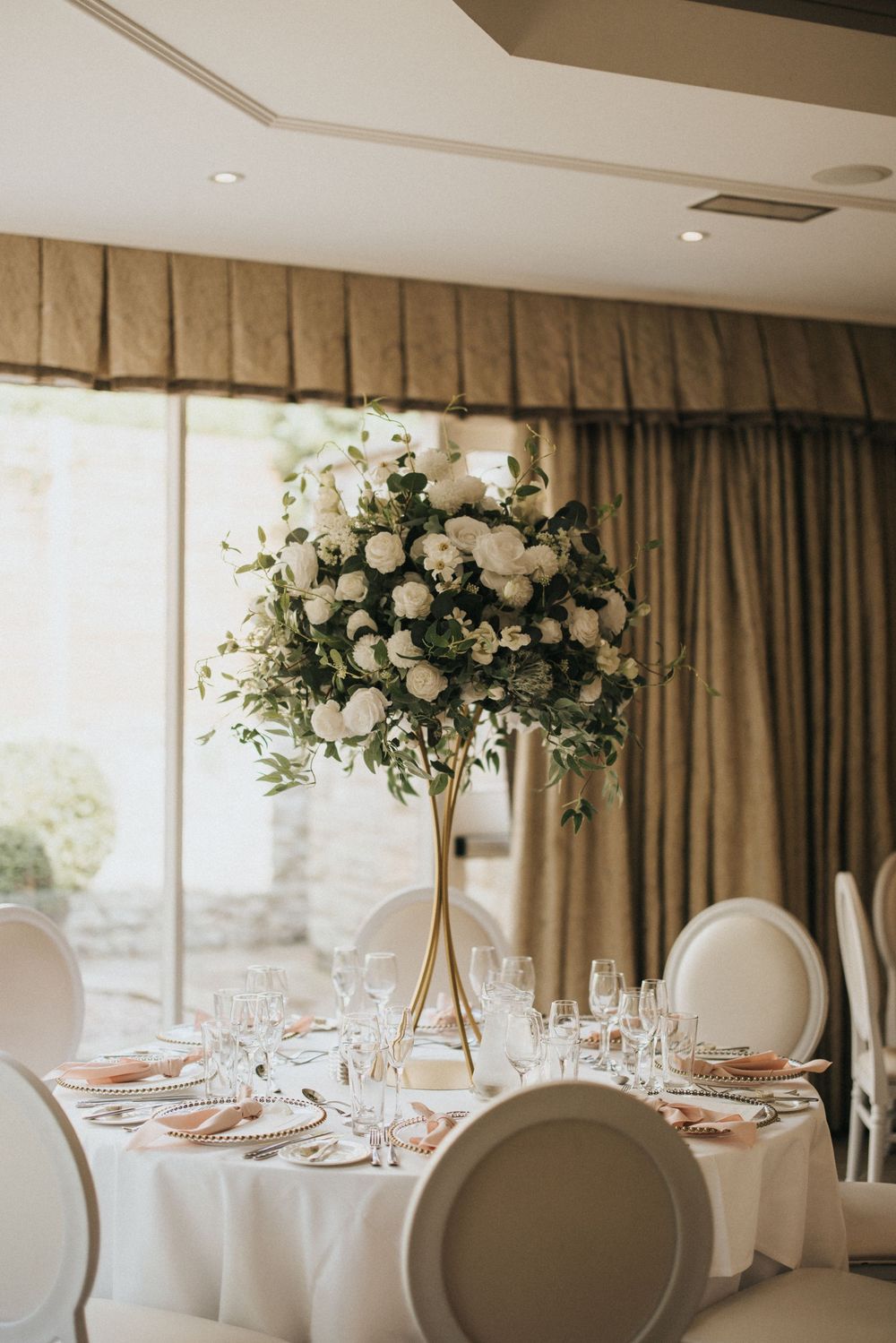 Elegant wedding reception table with tall white floral centerpiece, white linens, and cream-colored chairs in a formal banquet room.