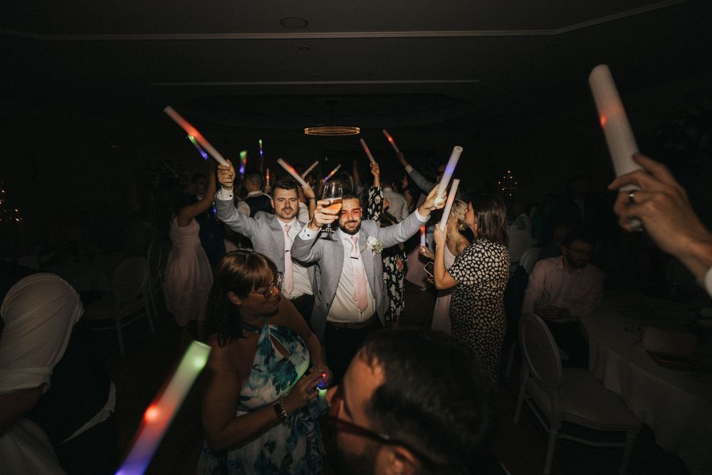 Party crowd dancing on a dark dance floor with glowsticks and hands raised in celebration during a nighttime event.