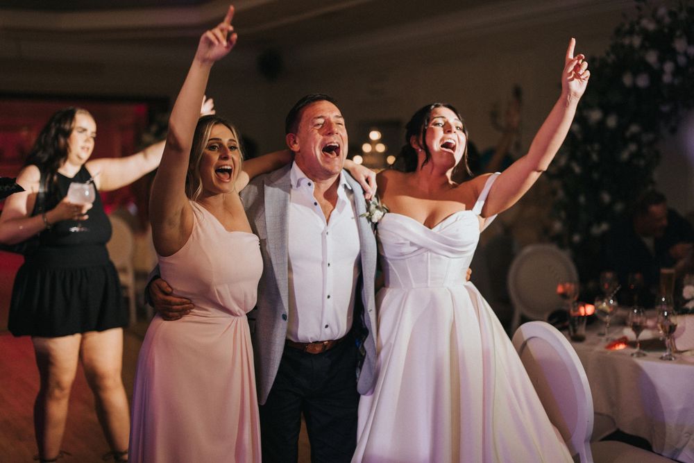 Lively wedding reception party guests singing and dancing with arms raised in celebration during evening festivities.