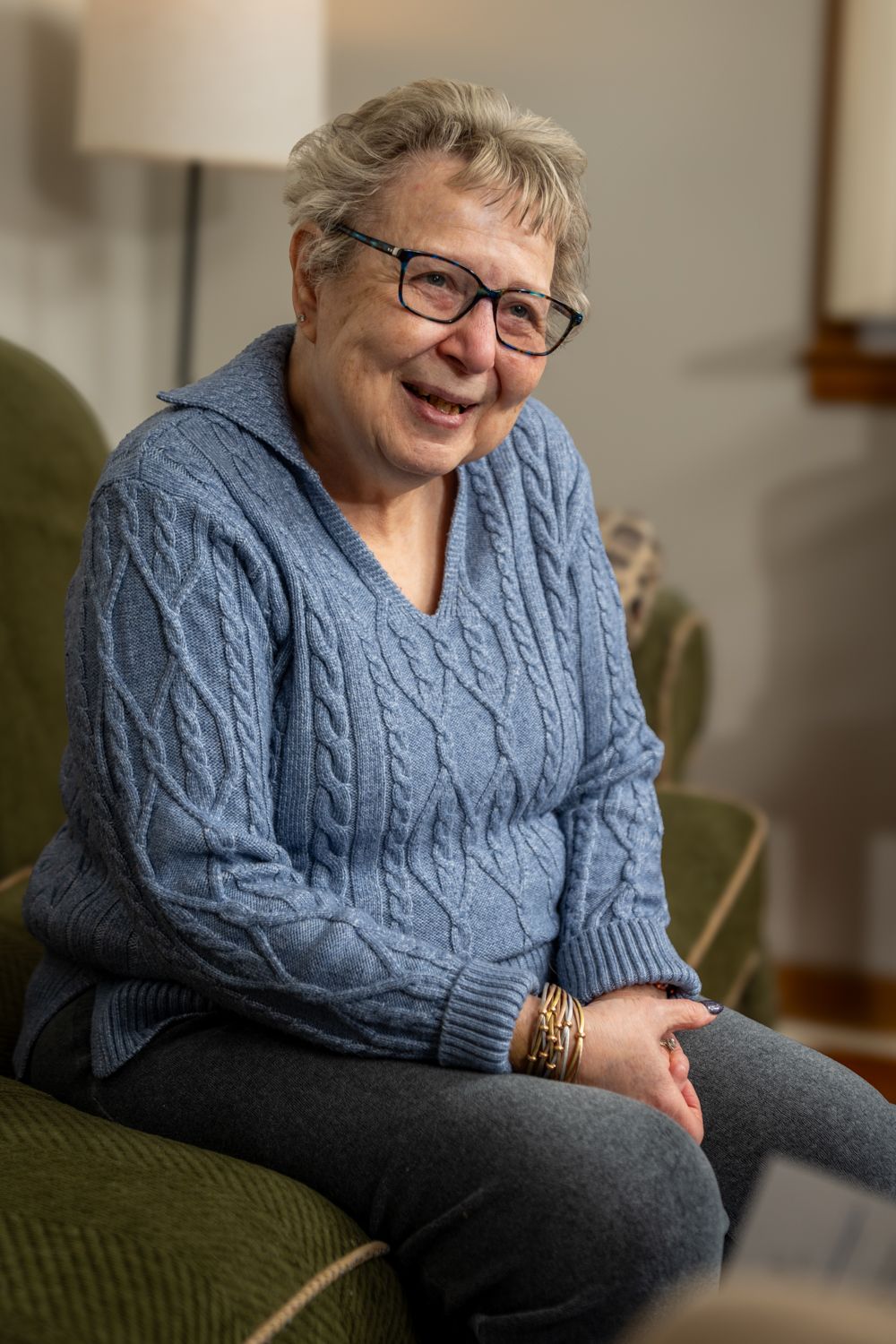 Smiling senior woman wearing blue cable knit sweater and glasses sitting casually on couch.