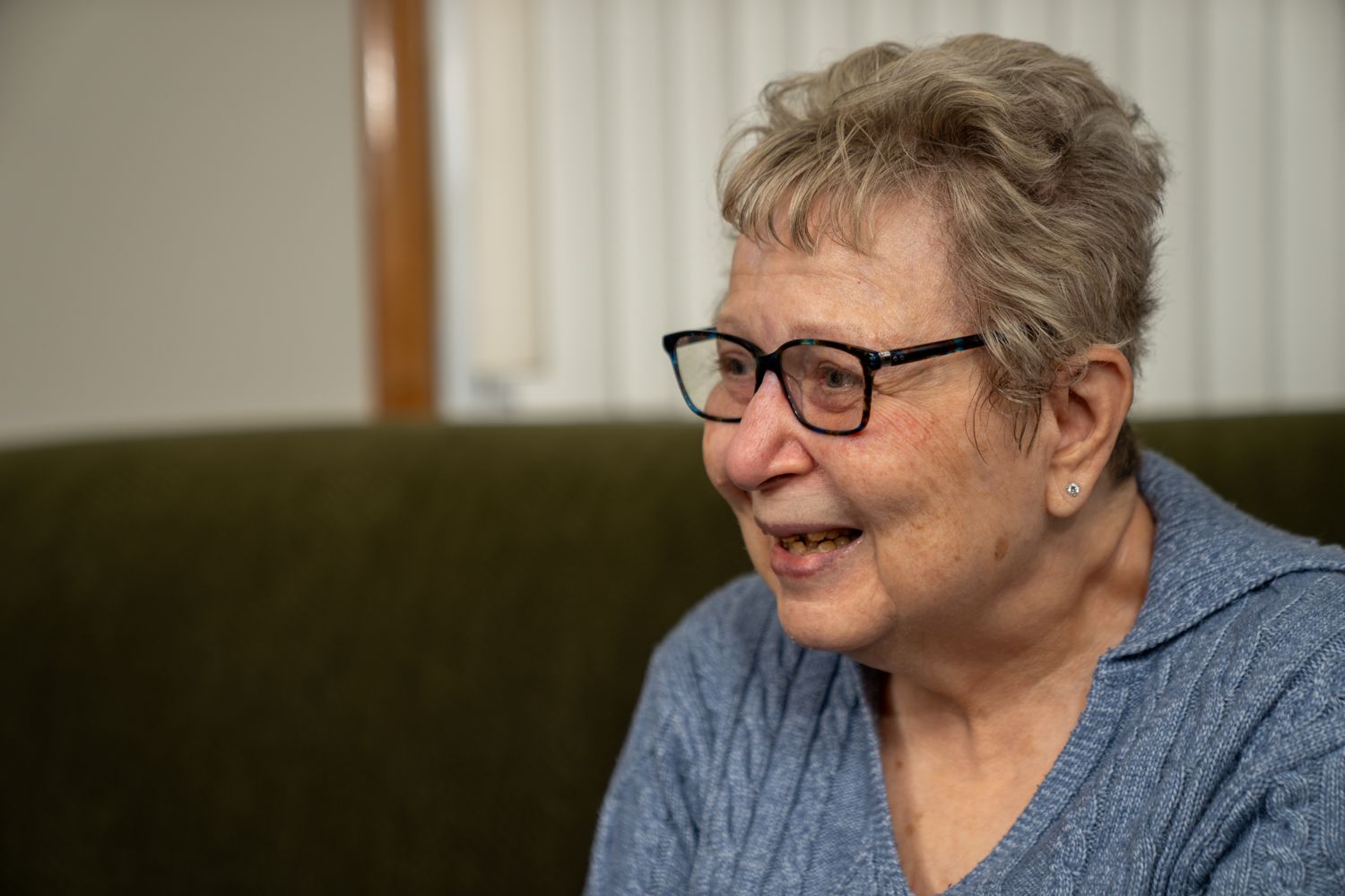 An elderly person wearing glasses and a blue sweater smiles warmly while seated indoors.