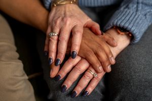 Close up photo of hands with sparkling jewelry rings and dark nail polish against gray knit fabric background.