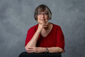 Portrait of a person in a bright red sweater with hand resting on chin in contemplative pose against gray background.