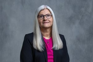 Professional portrait of someone with long silver hair wearing glasses, black blazer and bright pink top against gray backdrop.