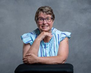 A person in glasses and blue ruffled blouse poses against grey backdrop with hand resting under their chin.
