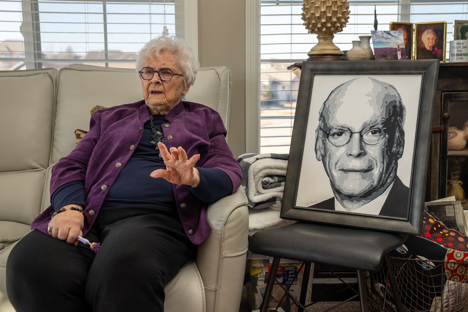 An elderly person wearing a purple sweater sits in an armchair reading a book in their home's living room setting.