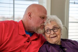 Tender moment between two people with one wearing red and one in purple by window.