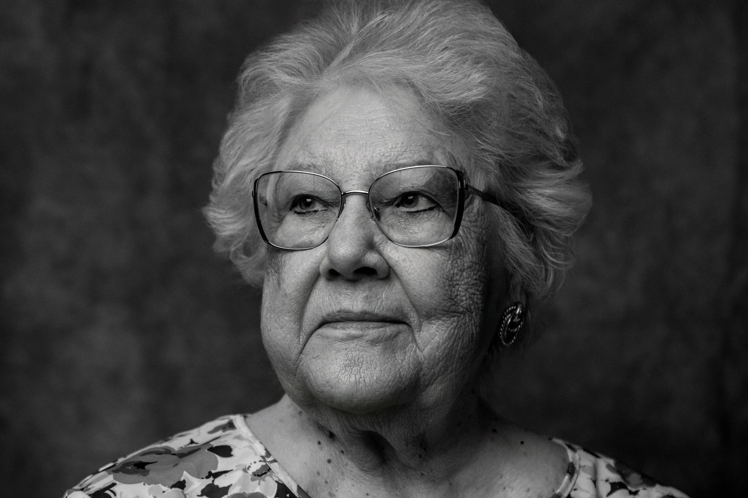 Black and white portrait of an elderly person wearing glasses and a floral print blouse against a dark background.