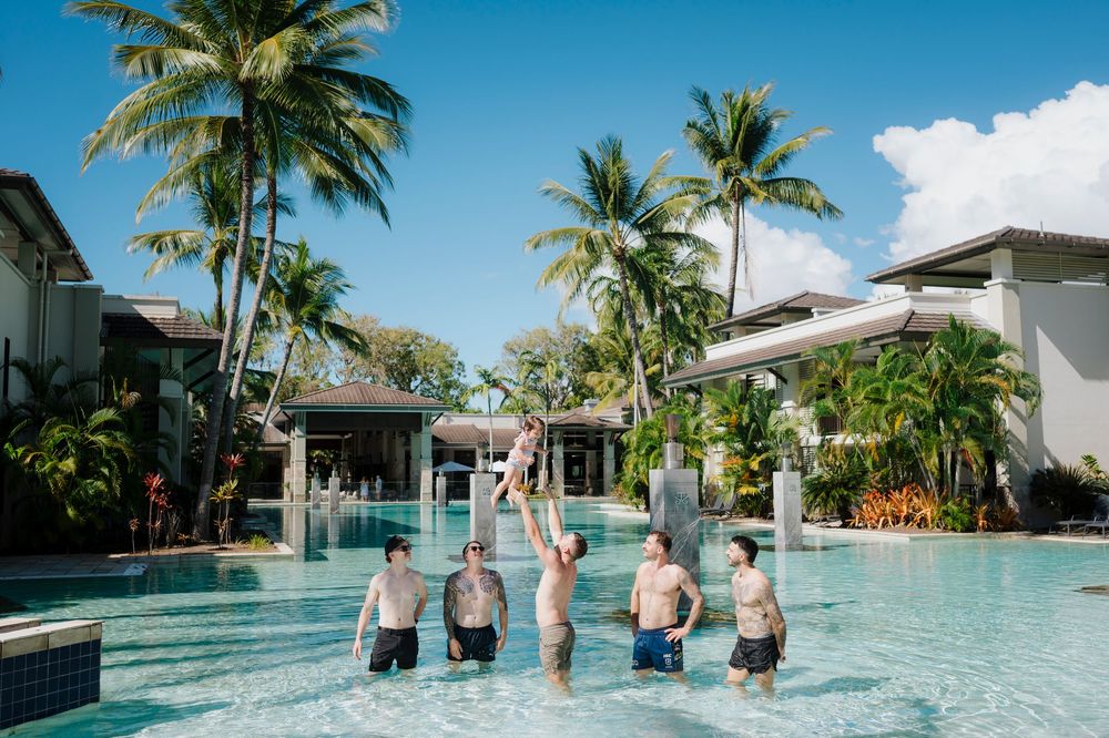 Group of friends playing and splashing in a tropical resort pool surrounded by palm trees.
