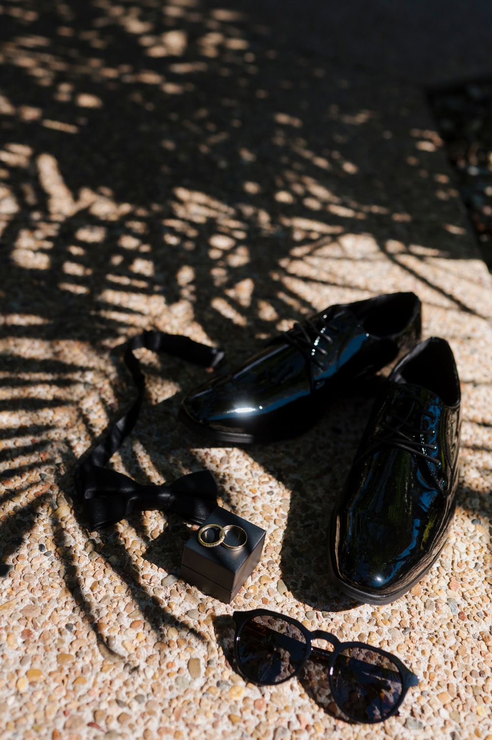 Wedding accessories including black dress shoes, rings and sunglasses casting dramatic shadows.