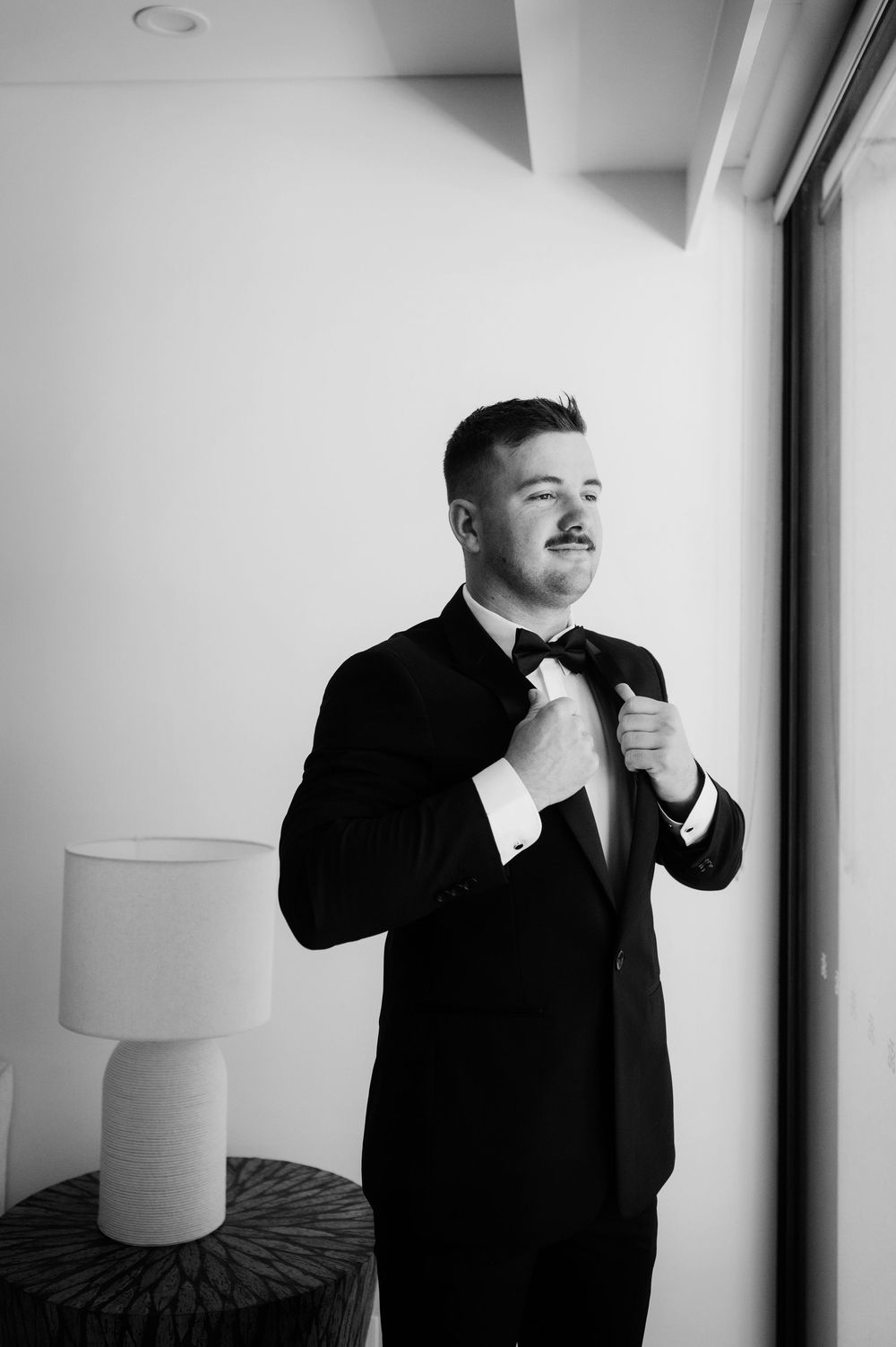 Classic black and white portrait of a person adjusting their bow tie by a window.