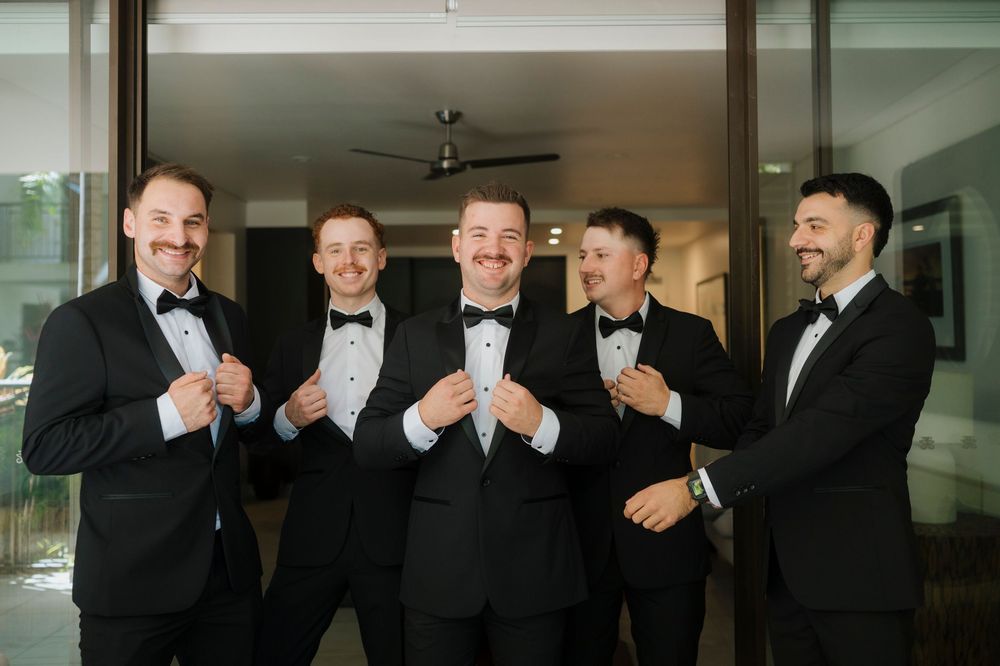 Groomsmen in black tuxedos sharing a lighthearted moment indoors.