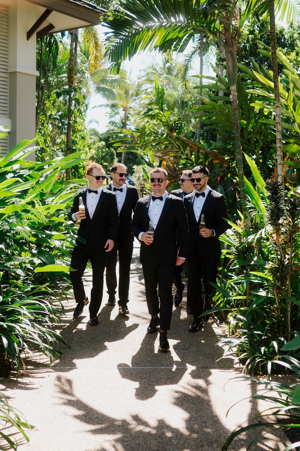 Wedding party in tuxedos walking through a lush tropical garden path.