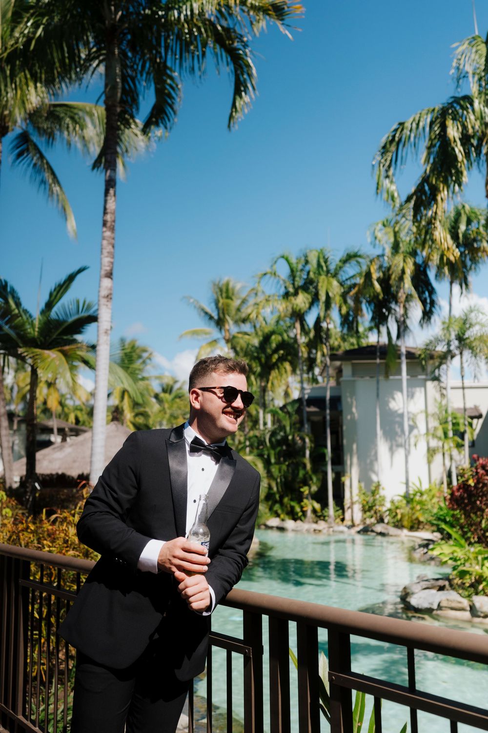 Person in a tuxedo with sunglasses enjoying a tropical poolside setting.