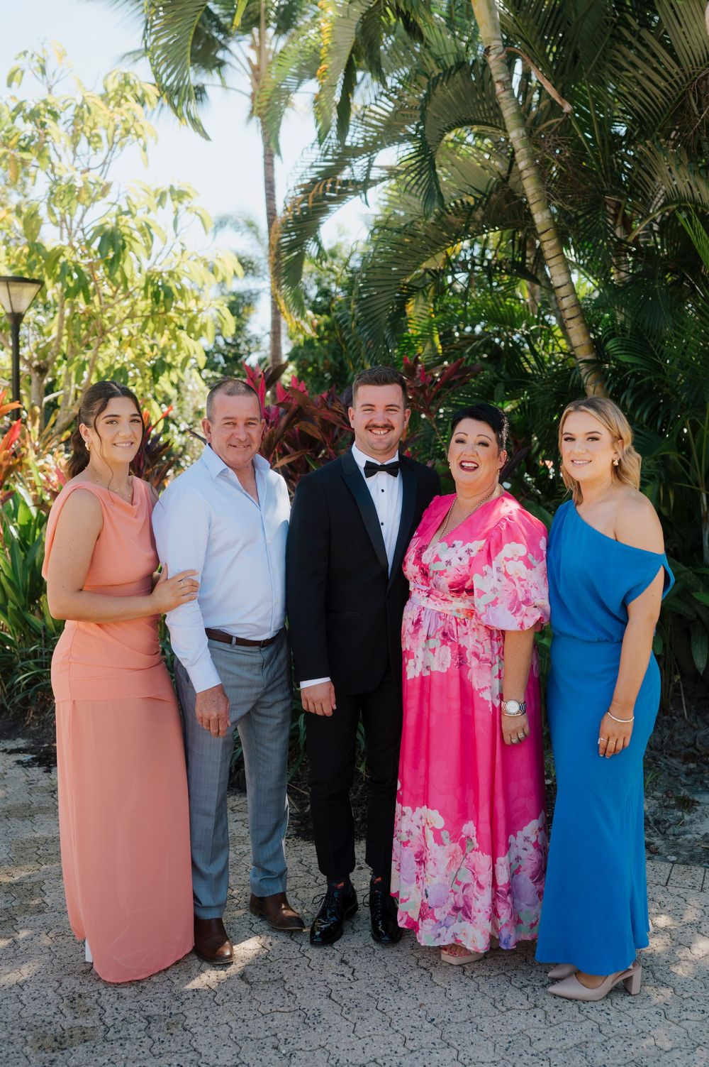 A formal group photo in a tropical garden setting with guests wearing colorful evening attire.