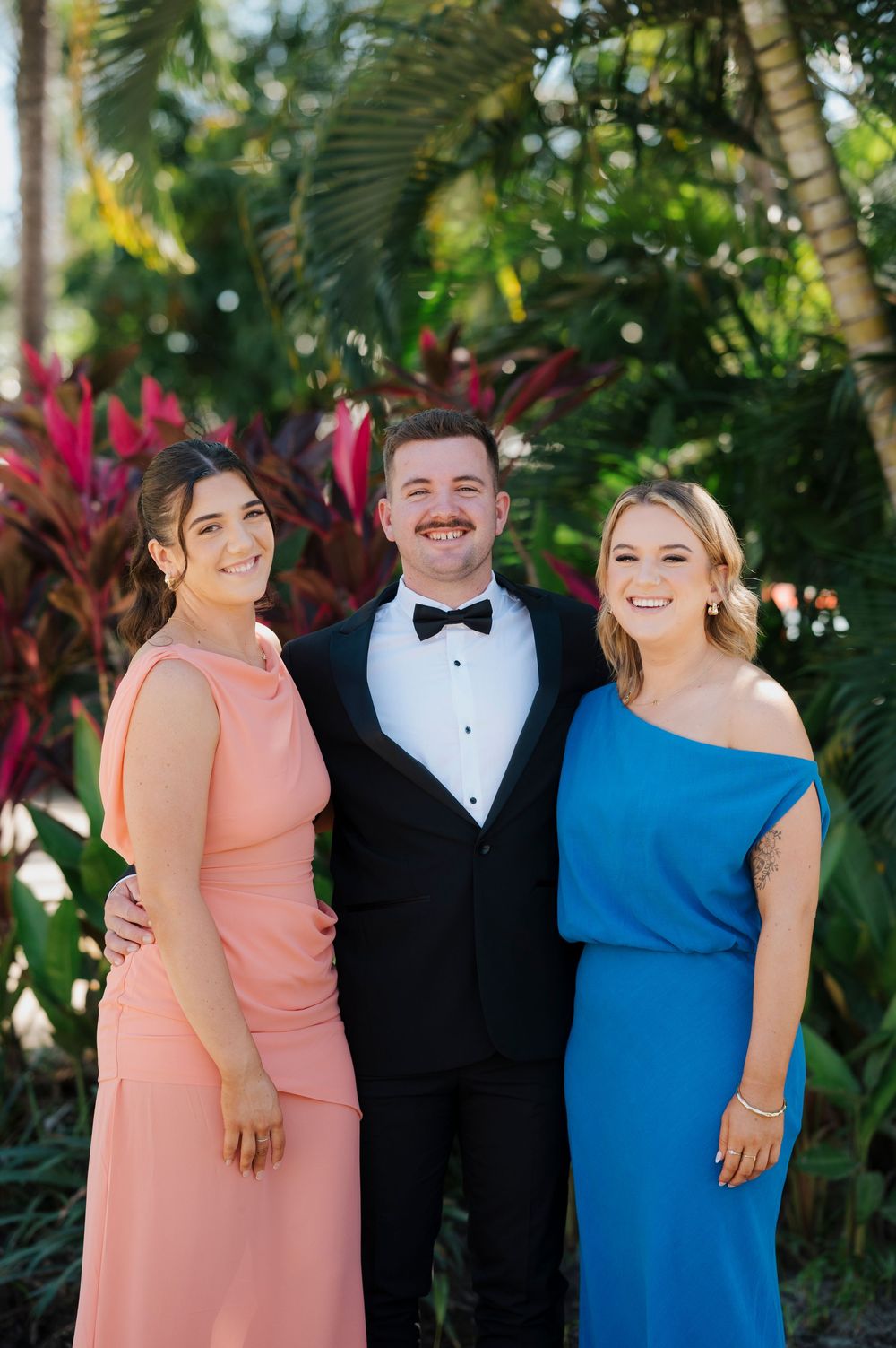 Three guests pose in formal attire with a tropical garden background.