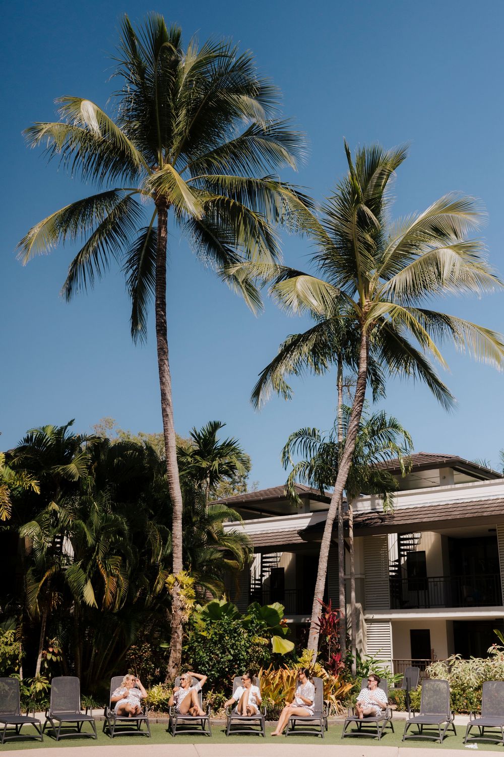 Palm trees and lounge chairs line the exterior of a resort building under blue sky.