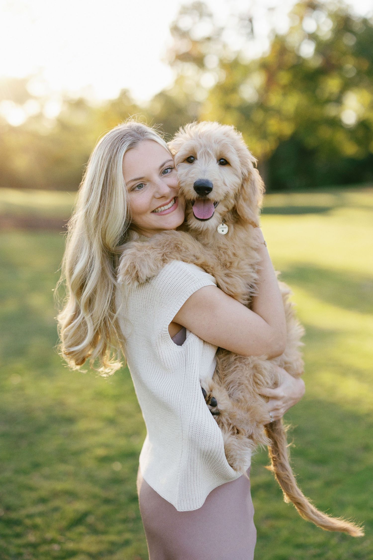 A happy Goldendoodle puppy being held outdoors during golden hour in a park setting.