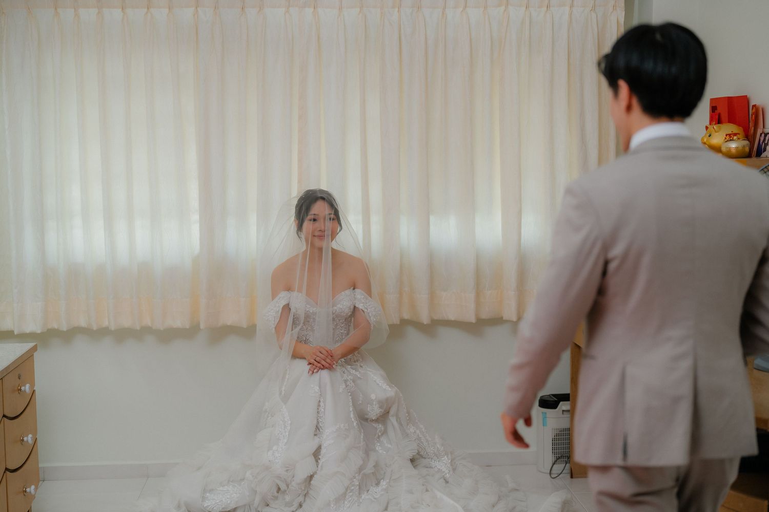 A bride in a flowing white gown sits in a sunlit room with soft curtains.
