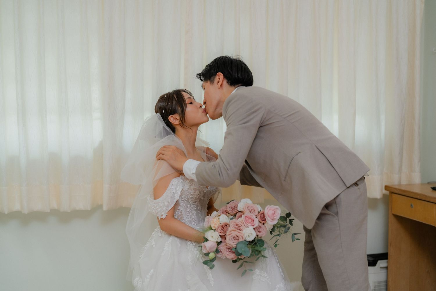A couple in wedding attire sharing an intimate moment against white curtains.