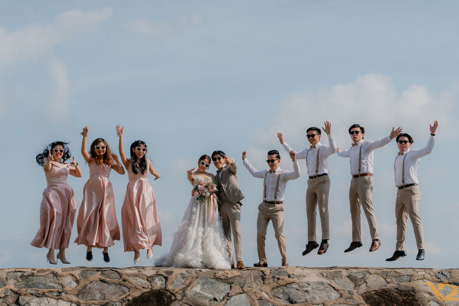 Wedding party jumping in synchronized pose on a stone wall against blue sky.