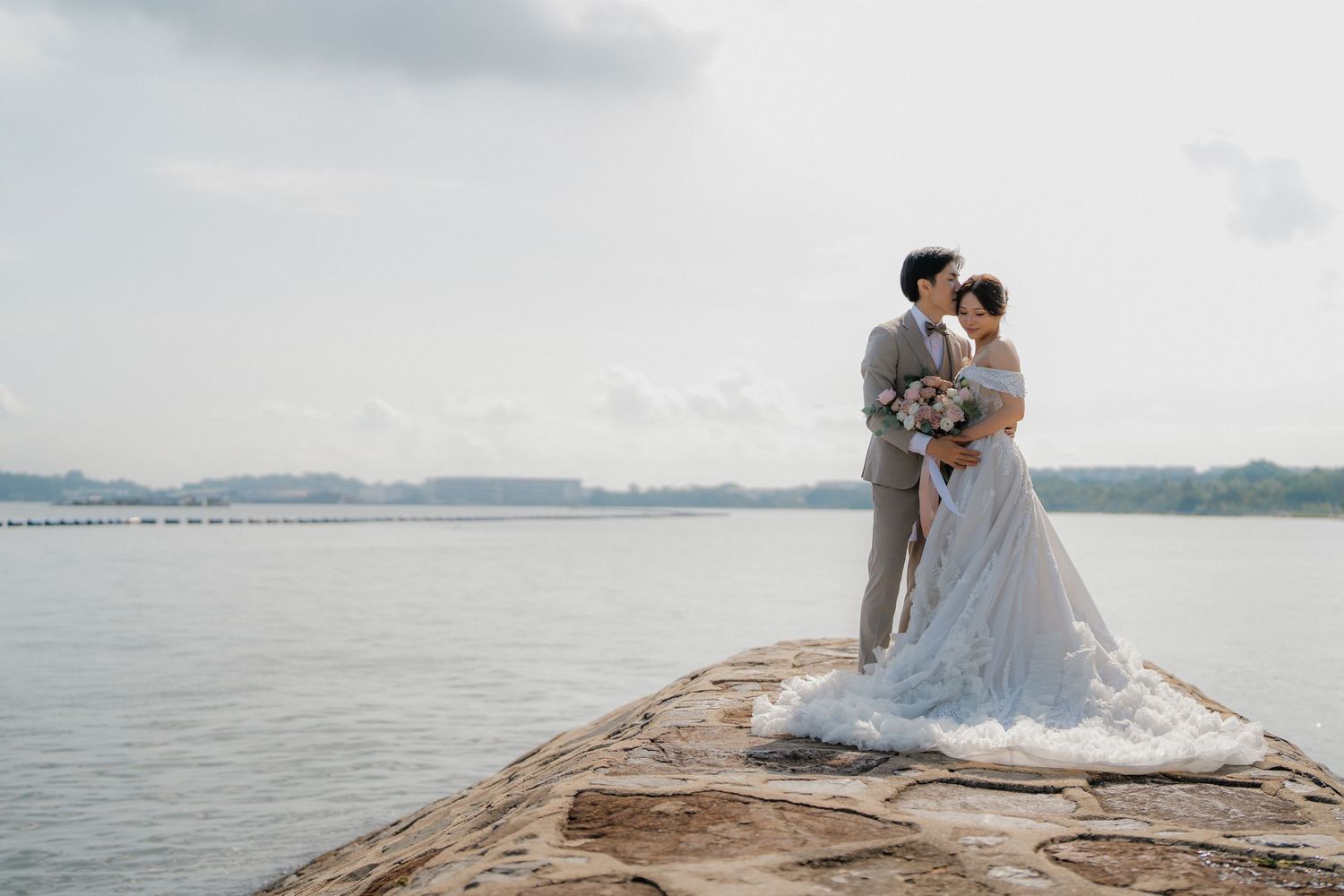 A couple stands on a rocky outcrop overlooking a serene lake during their wedding photoshoot.