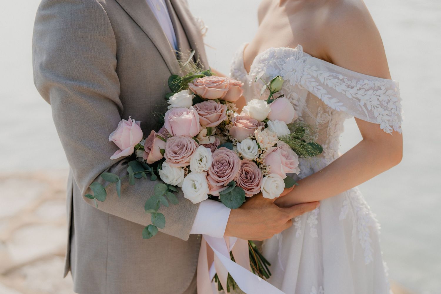 A couple holding a romantic wedding bouquet with pink roses and greenery against a light background.