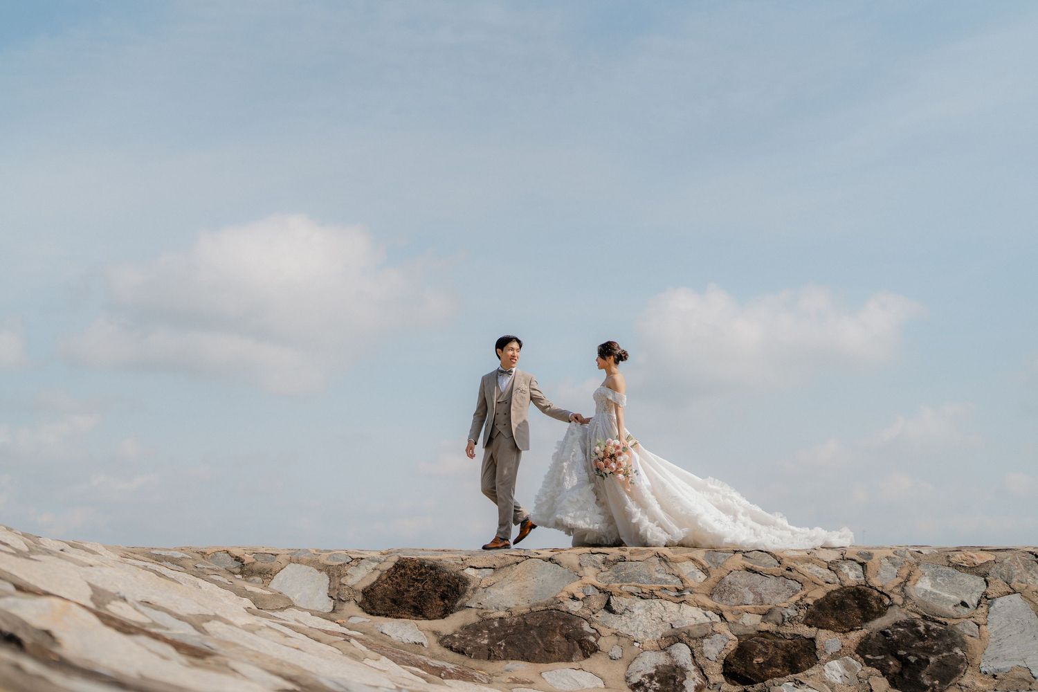 A couple walks along a rocky seaside landscape in wedding attire during a photo session.